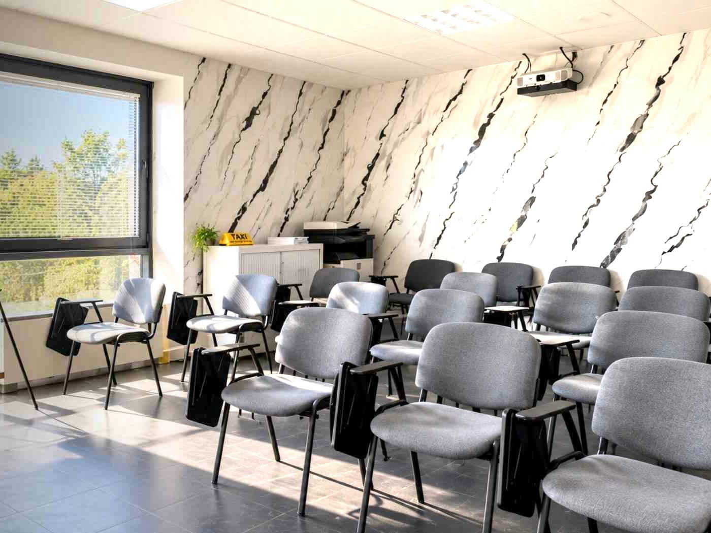 La salle de cours de BIG FORMA : rangées de chaises à tablette, vidéoprojecteur au plafond, lumineux TAXI sur le meuble et grande baie vitrée.